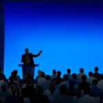 Man at a lectern in front of a crowd, big blue screen behind him