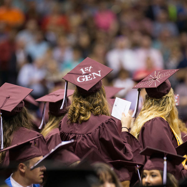 A sea of students at graduation with maroon caps and gowns. One cap says 1st Gen on it.