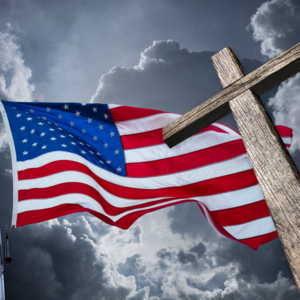 A Christian cross rises imposingly into frame, while in the background a U.S. flag waves against dark clouds